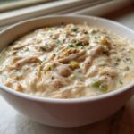 A close-up of a white bowl filled with creamy, shredded chicken chili, garnished with herbs.