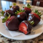 A plate of glossy chocolate covered strawberries, one cut in half showing the red interior.