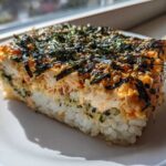 A close-up slice of baked rice, creamy topping, and salmon, garnished with shredded nori and sesame seeds for sushi bake.
