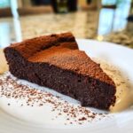 A decadent slice of flourless chocolate torte dusted heavily with cocoa powder, served on a white plate.