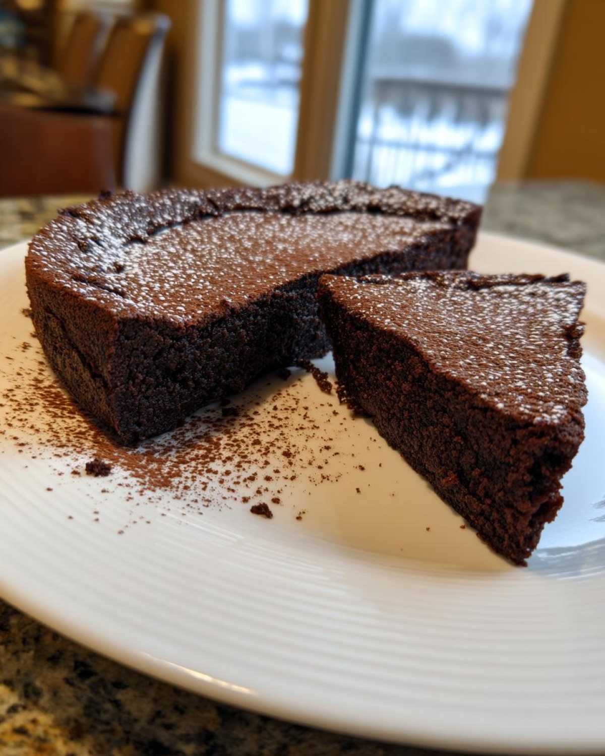 A rich, dark slice cut from a flourless chocolate torte, dusted with powdered sugar on a white plate.