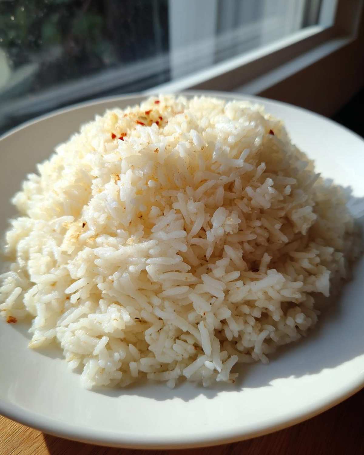 A mound of fluffy white butter rice served on a white plate, illuminated by bright sunlight.