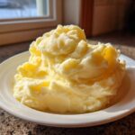 A generous serving of creamy, fluffy mashed potatoes piled high on a white bowl, highlighted by sunlight.