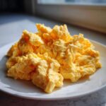 A generous serving of bright yellow, fluffy scrambled eggs piled high on a white plate near a window.