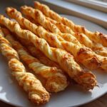 Close-up of several golden brown, twisted cheese straws recipe baked on a white plate near a window.