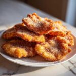 A stack of freshly fried, golden buñuelos generously coated in cinnamon sugar on a white plate.