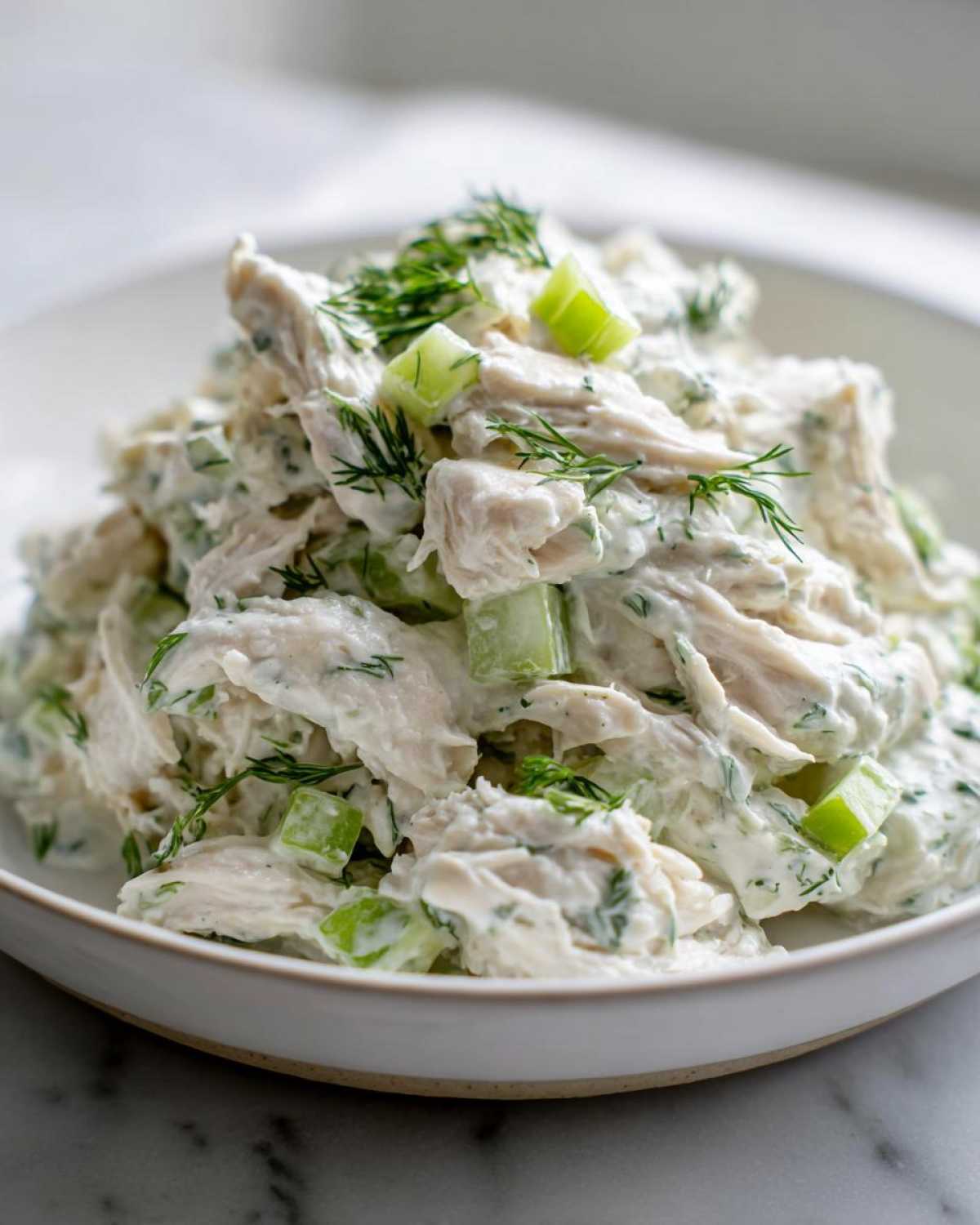 Close-up of shredded chicken mixed with a creamy dressing, celery chunks, and fresh dill, representing greek yogurt chicken salad.