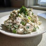 A mound of creamy greek yogurt chicken salad mixed with celery and topped with fresh dill, served on a white plate.