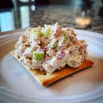 A scoop of creamy ham salad recipe with celery and red onion served on a square cracker on a white plate.