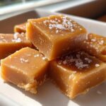 Close-up of chewy homemade caramel squares sprinkled with flaky sea salt on a white plate.