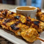 Close-up of perfectly grilled, golden-brown chicken satay skewers served with a side of peanut dipping sauce.