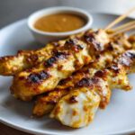 Close-up of perfectly grilled chicken satay skewers served with a side of creamy peanut dipping sauce.