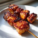 Close-up of two skewers holding pieces of juicy, glazed, and slightly charred chicken skewers on a white plate.