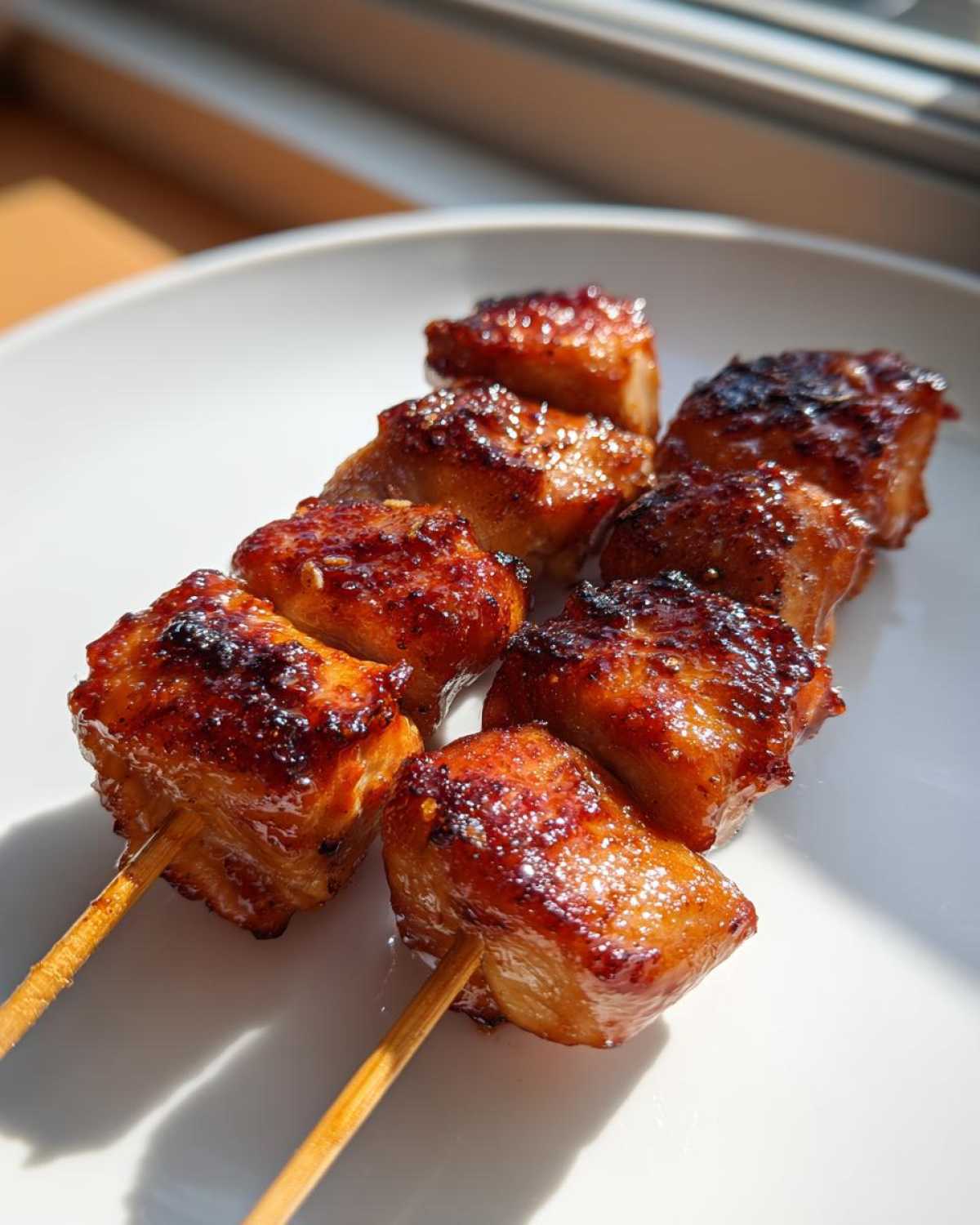 Two wooden sticks holding glistening, caramelized chicken skewers resting on a white plate.