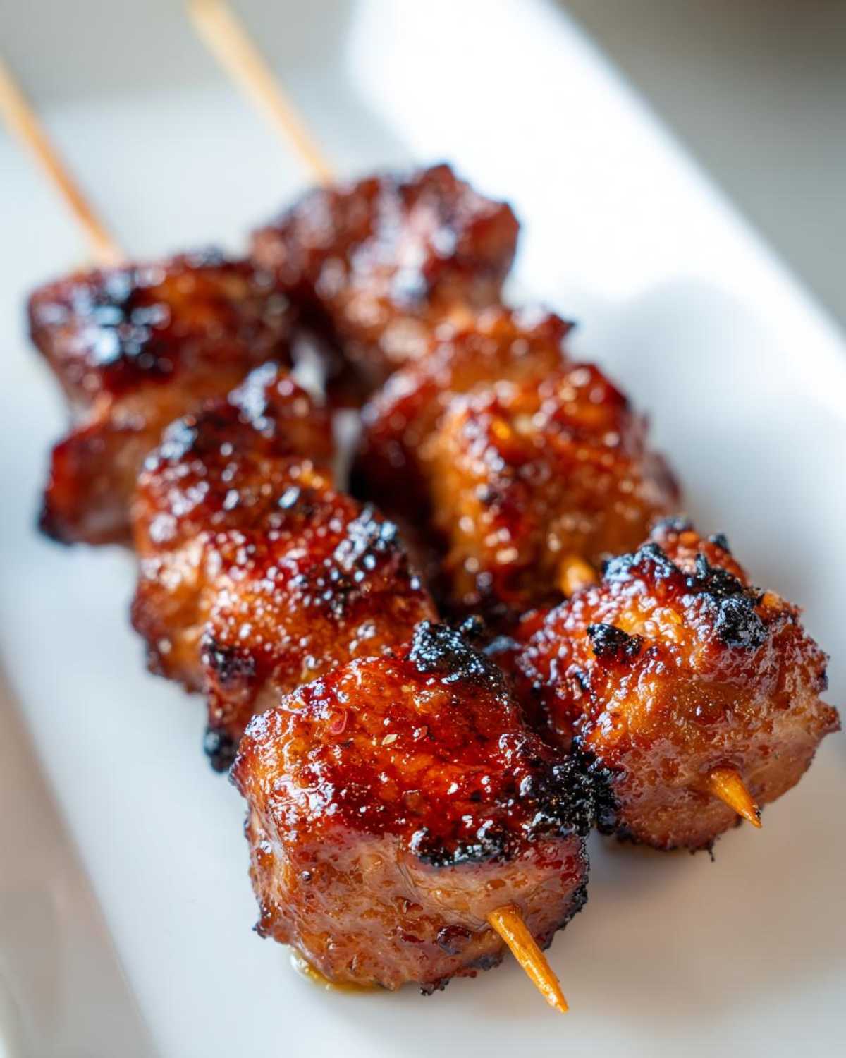 Close-up of several pieces of glazed, charred chicken skewers resting on a white plate.