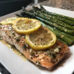 A perfectly cooked salmon recipe fillet topped with lemon slices and herbs, served with roasted asparagus.