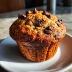 A golden brown banana chocolate chip muffin topped with melted chocolate chips, sitting on a small white plate.