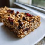 A close-up of a thick, chewy no bake granola bars piece topped with chocolate chips and dried cranberries.
