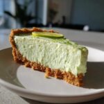 A thick slice of creamy, pale green no-bake key lime cheesecake with a graham cracker crust, topped with lime zest and a lime slice.