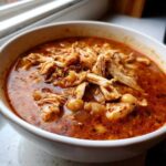 Close-up of a white bowl filled with rich, red chicken pozole, featuring shredded chicken and hominy kernels.