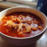 Close-up of a white bowl filled with rich, red broth chicken pozole, featuring shredded chicken and hominy.
