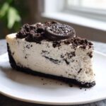 A decadent slice of oreo cheesecake with a thick dark crust, creamy filling mixed with cookie pieces, and topped with crumbs and a whole Oreo.