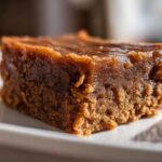 A close-up side view of a single, rich brown square of homemade penuche fudge resting on a white plate.