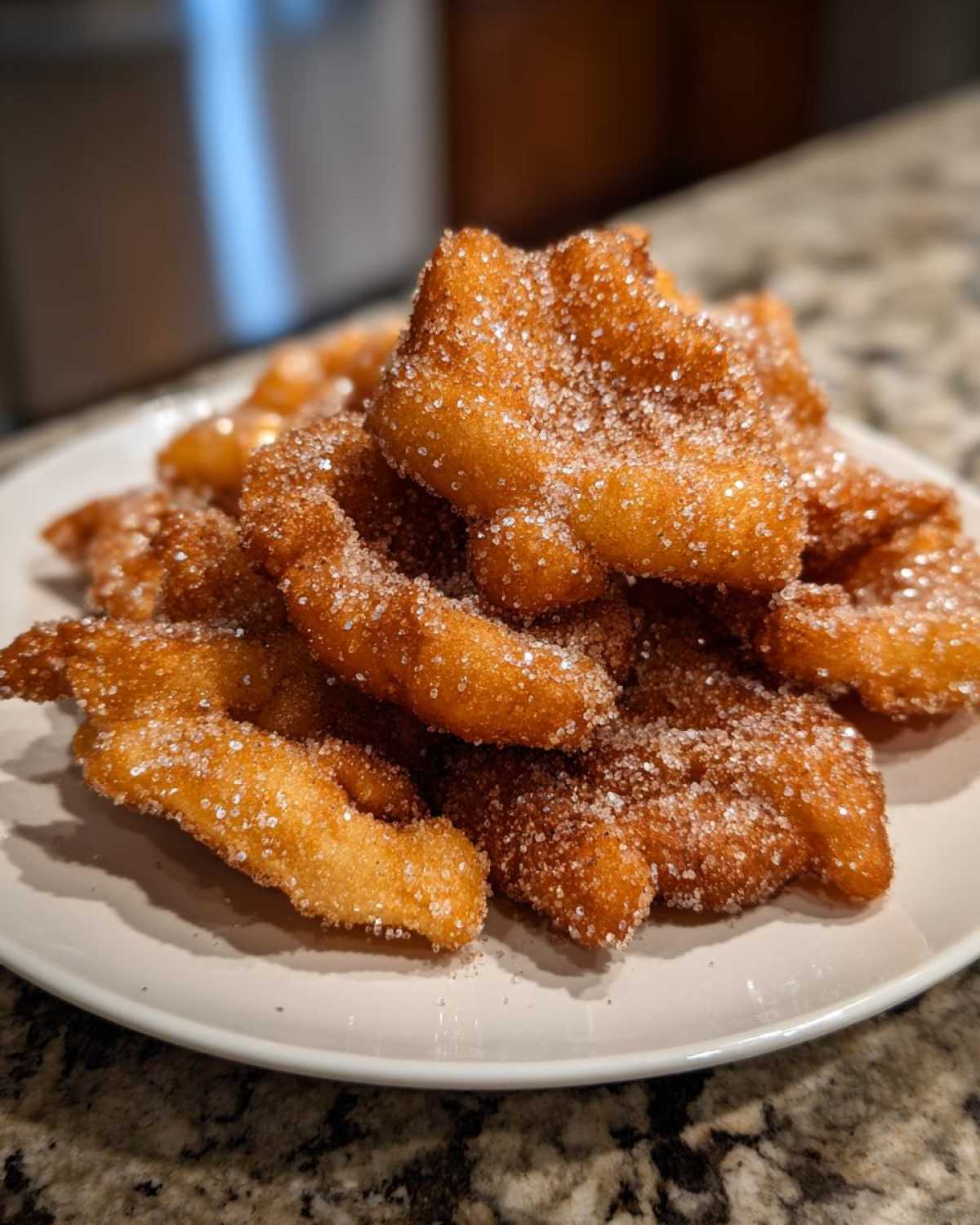 A plate piled high with freshly fried, golden brown buñuelos heavily coated in sparkling sugar.