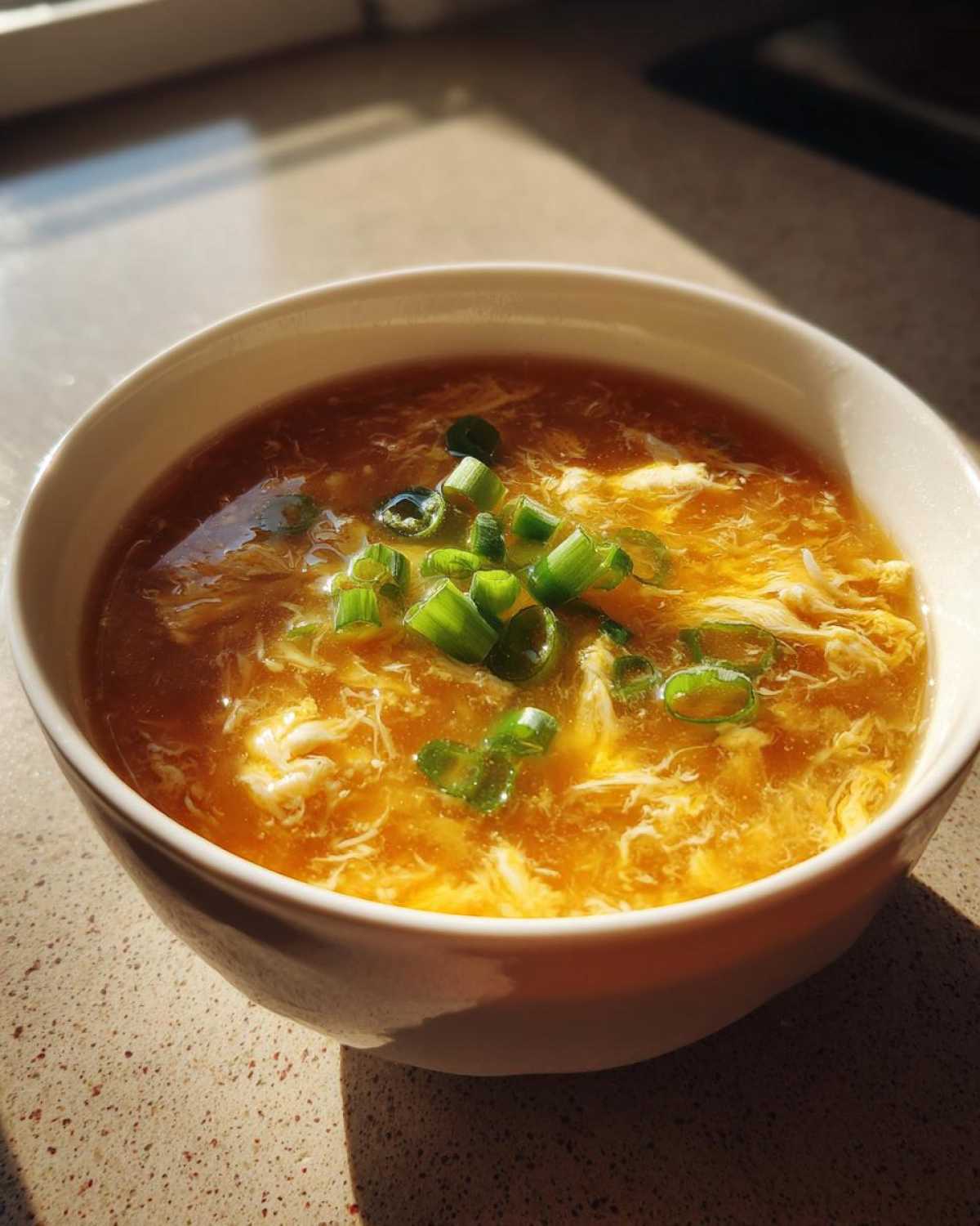 A close-up of a white bowl filled with golden yellow egg drop soup, garnished with chopped green onions.
