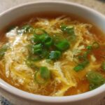 A close-up view of a bowl of hot egg drop soup featuring wispy egg ribbons and fresh green onion garnish.