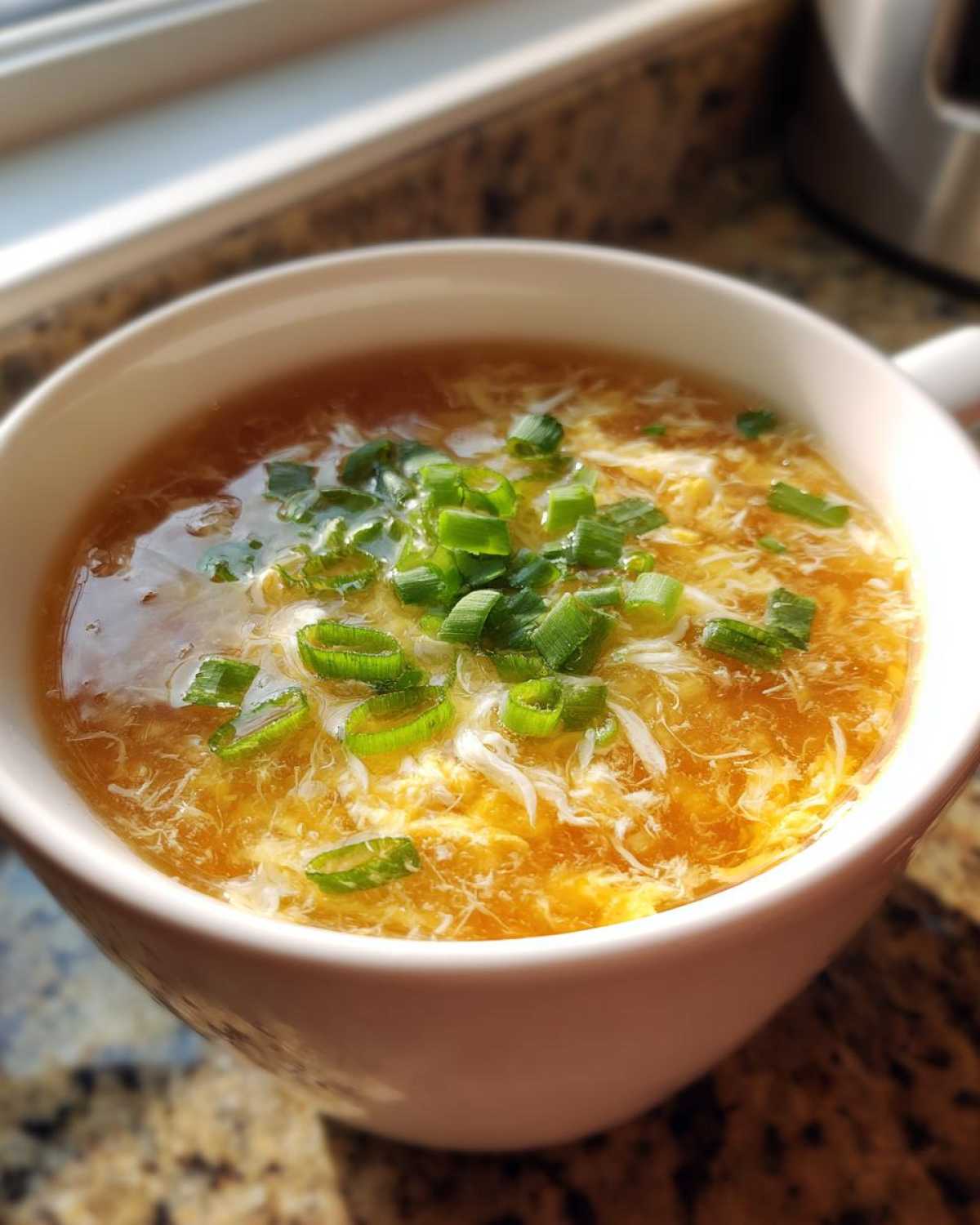 A close-up of a white mug filled with hot egg drop soup, garnished with fresh green scallions.