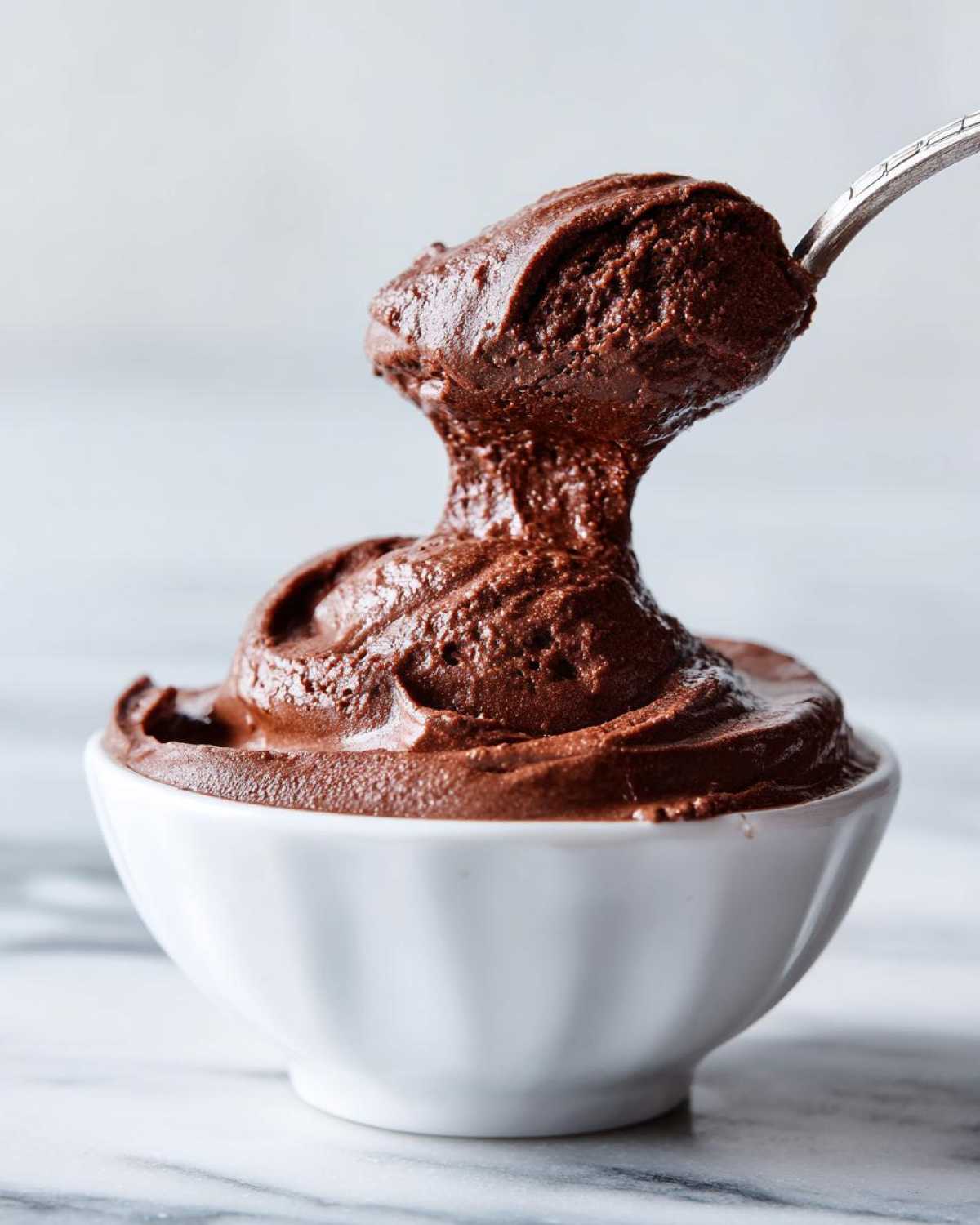 A spoonful of rich, dark chocolate mousse is lifted from a small white bowl, showing its thick texture.