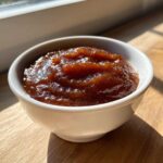 A close-up of thick, dark brown pear butter glistening in a small white bowl on a wooden surface.