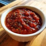 Close-up of thick, rich red pizza sauce seasoned with dried herbs, served in a small white ramekin.