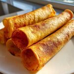 Close-up of golden, rolled tortillas, likely the base for potato tacos, stacked on a white plate.