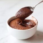 A spoonful of rich, airy chocolate mousse being lifted from a white bowl dusted with cocoa powder.