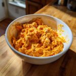 A white bowl filled with bright orange, shredded buffalo chicken mixture sitting on a wooden cutting board.