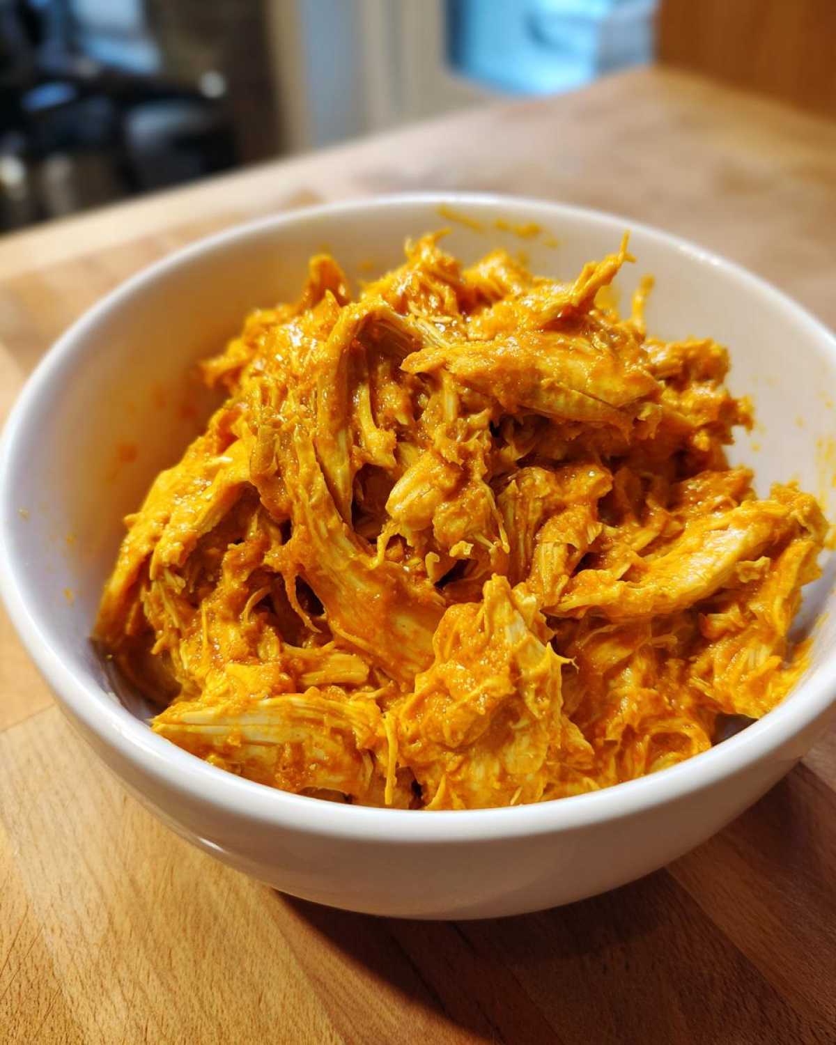 Close-up of shredded buffalo chicken coated in a vibrant orange sauce, served in a white bowl.