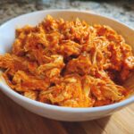 Close-up of shredded buffalo chicken coated in bright orange sauce, served in a white bowl.