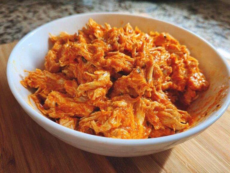 Close-up of shredded buffalo chicken coated in bright orange sauce, served in a white bowl.