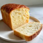 A soft, fluffy loaf of condensed milk bread with one slice cut and resting beside it on a white plate.