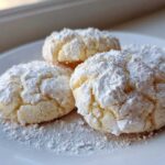 A close-up of four soft, crinkle-top cool whip cookies heavily dusted with powdered sugar on a white plate.