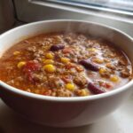 A close-up of a steaming white bowl filled with rich cowboy soup containing ground beef, corn, and kidney beans.