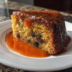 A close-up of a moist slice of sticky toffee pudding recipe covered in rich toffee sauce on a white plate.