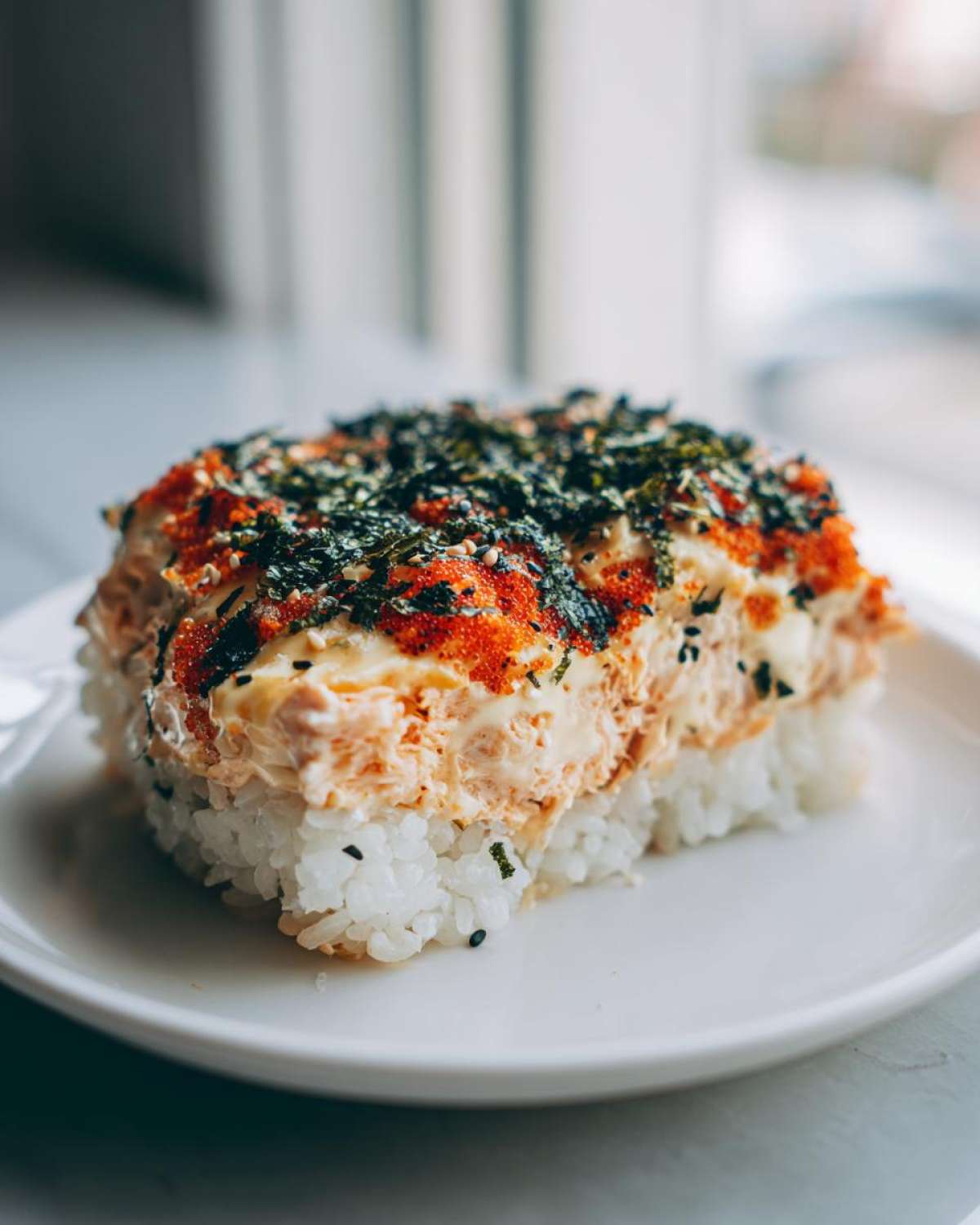 A close-up serving of baked sushi layered over rice, topped with creamy sauce, bright orange masago, and shredded nori.