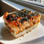 A square slice of baked salmon sushi bake topped with furikake and sesame seeds on a white plate.