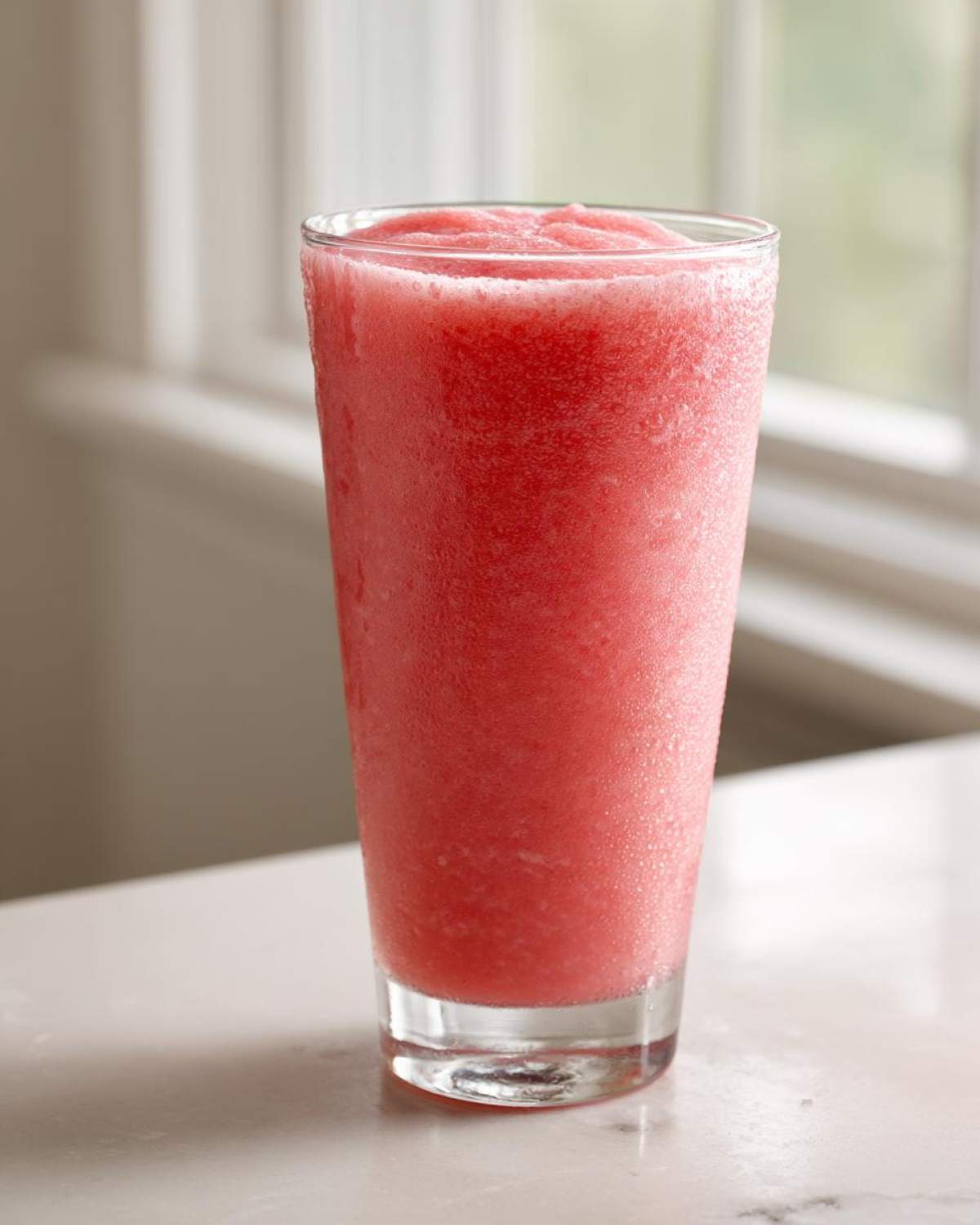 A tall glass filled with a frosty, bright pink watermelon smoothie sitting on a white counter.