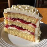 A thick slice of white chocolate raspberry cake showing three layers of yellow cake, raspberry filling, and white frosting.