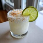 A creamy white Christmas margarita garnished with toasted coconut flakes and a fresh lime wheel.