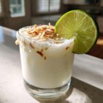 A creamy white Christmas margarita garnished with toasted coconut flakes and a fresh lime wheel.